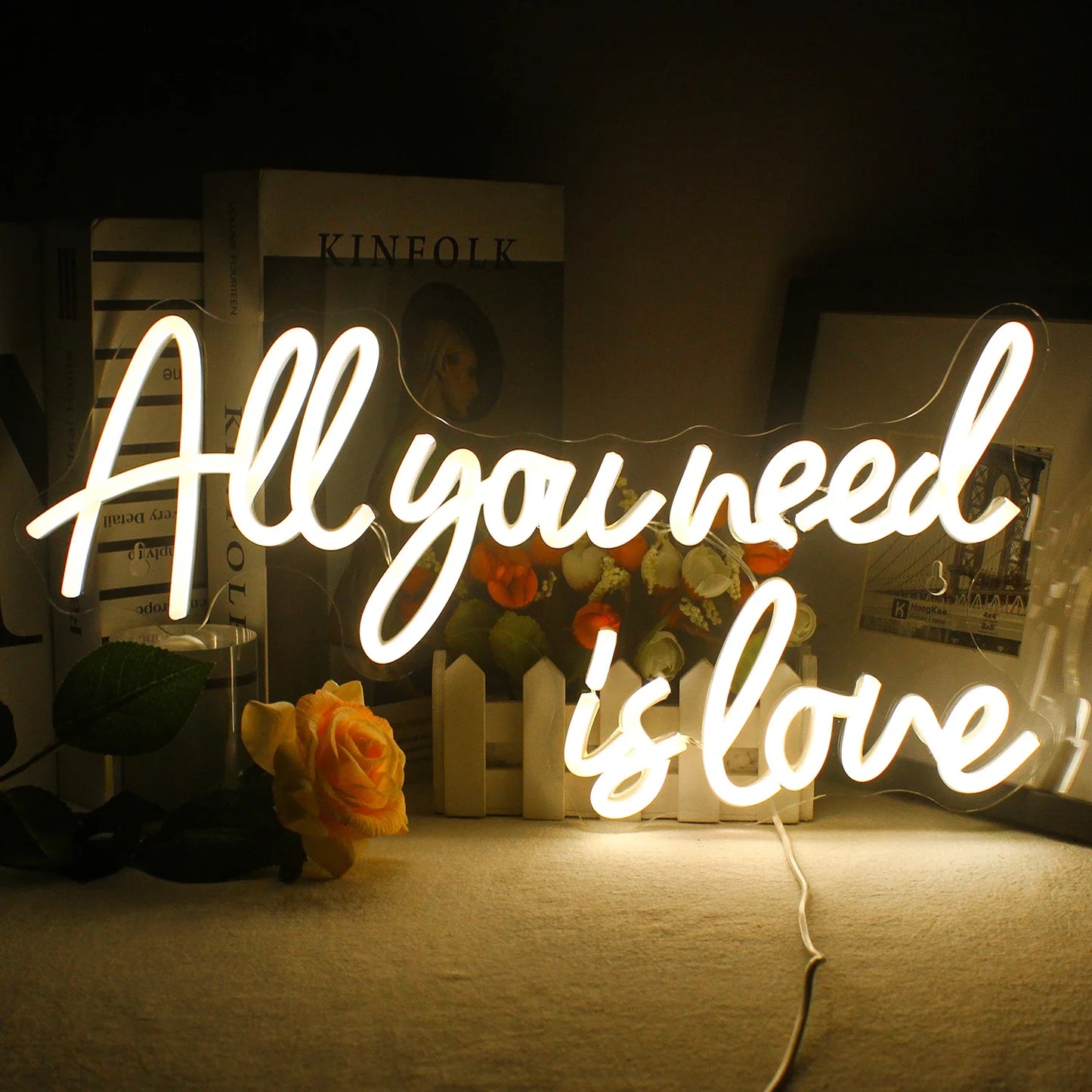 Glowing neon sign reading "All you need is love" in cursive, placed on a table with a rose, flowers, and books in the background, creating a cozy atmosphere.
