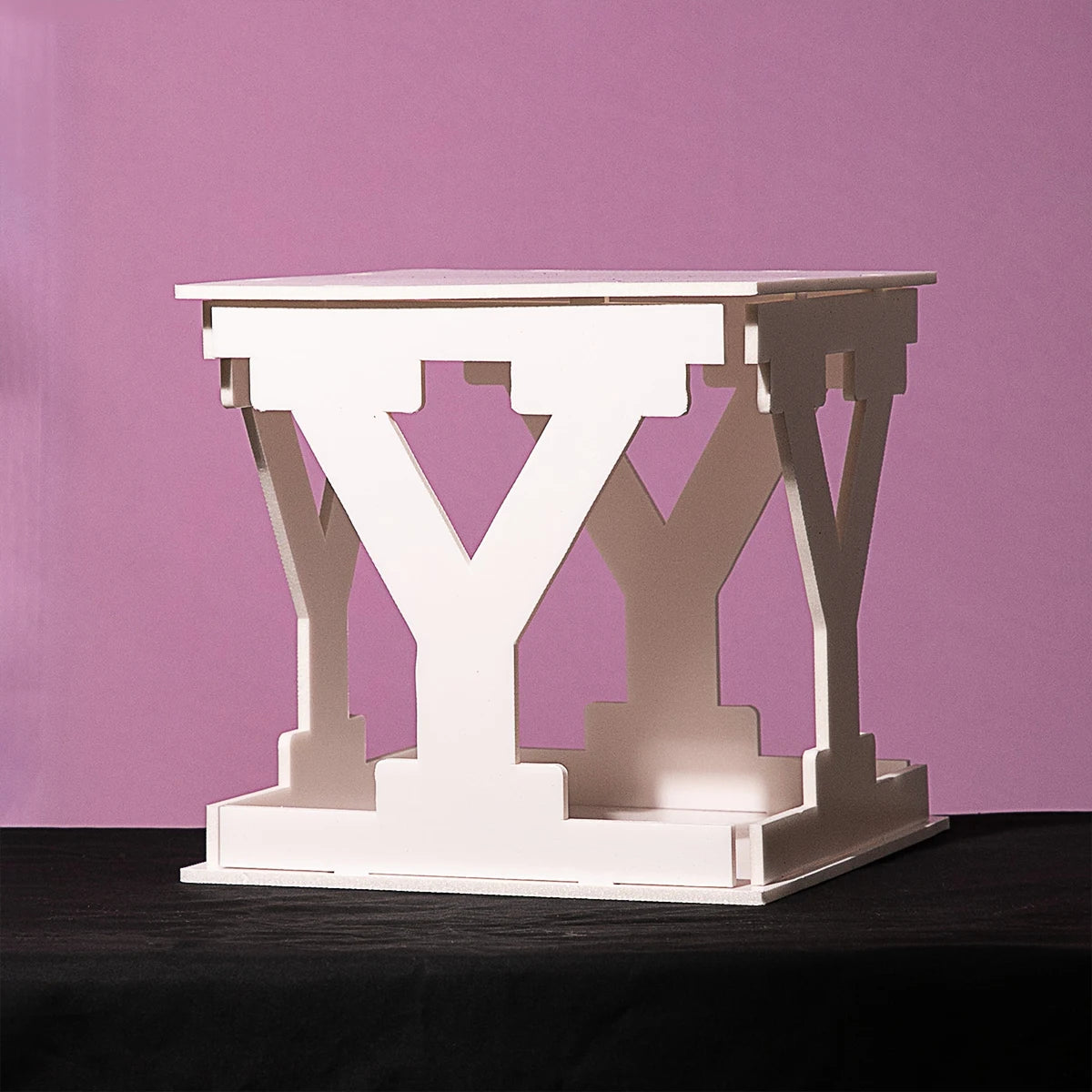 White geometric Y-shaped table on a black surface with a pastel purple background. The table has a minimalist design with symmetrical supports and a flat square top.