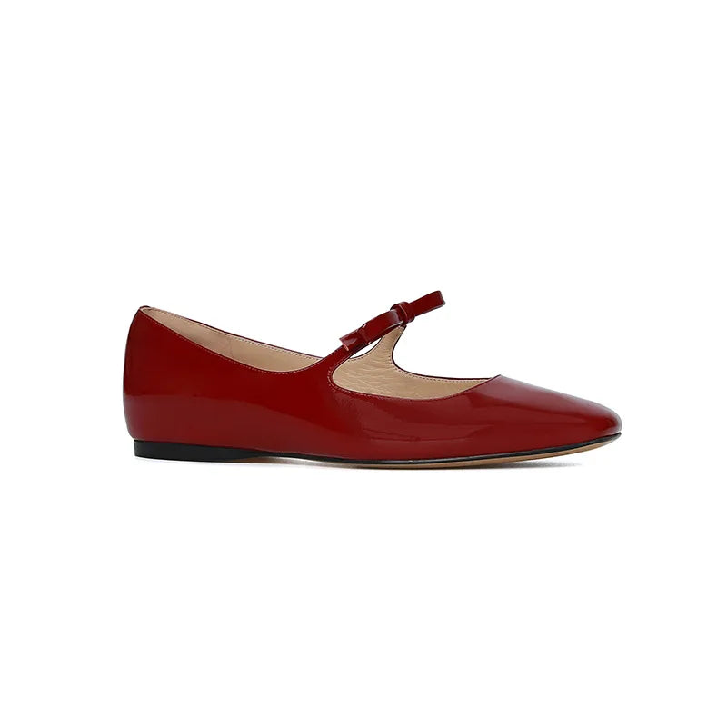 Red patent leather Mary Jane flat shoe with a single strap and bow detail on a low black sole. Minimalist and elegant design.