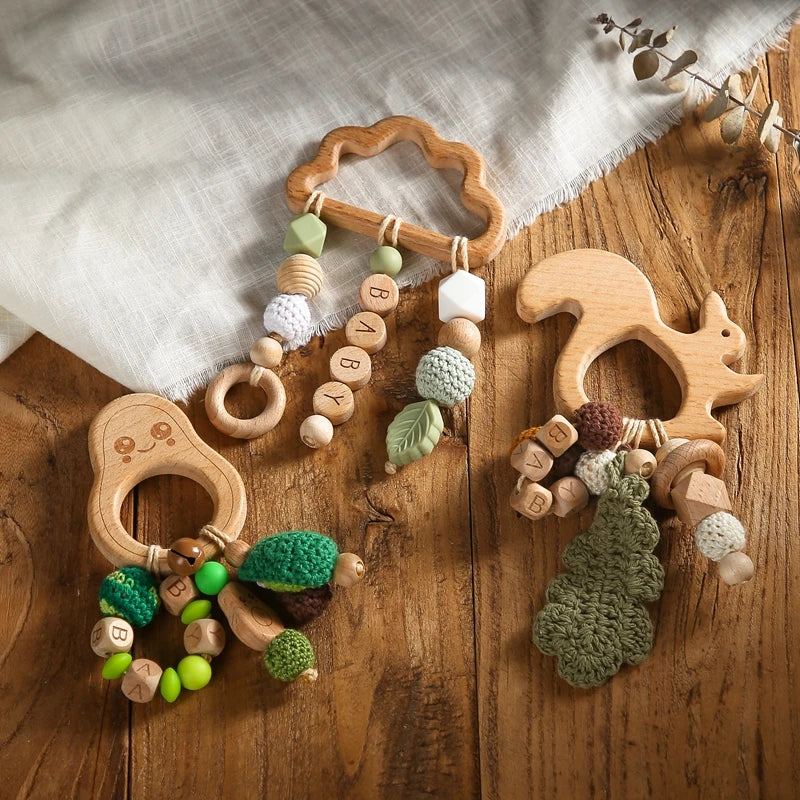 Wooden baby teethers in cloud, bear, and squirrel shapes with knitted and wooden bead ornaments, labeled "BABY." Placed on a wooden surface with a fabric backdrop.