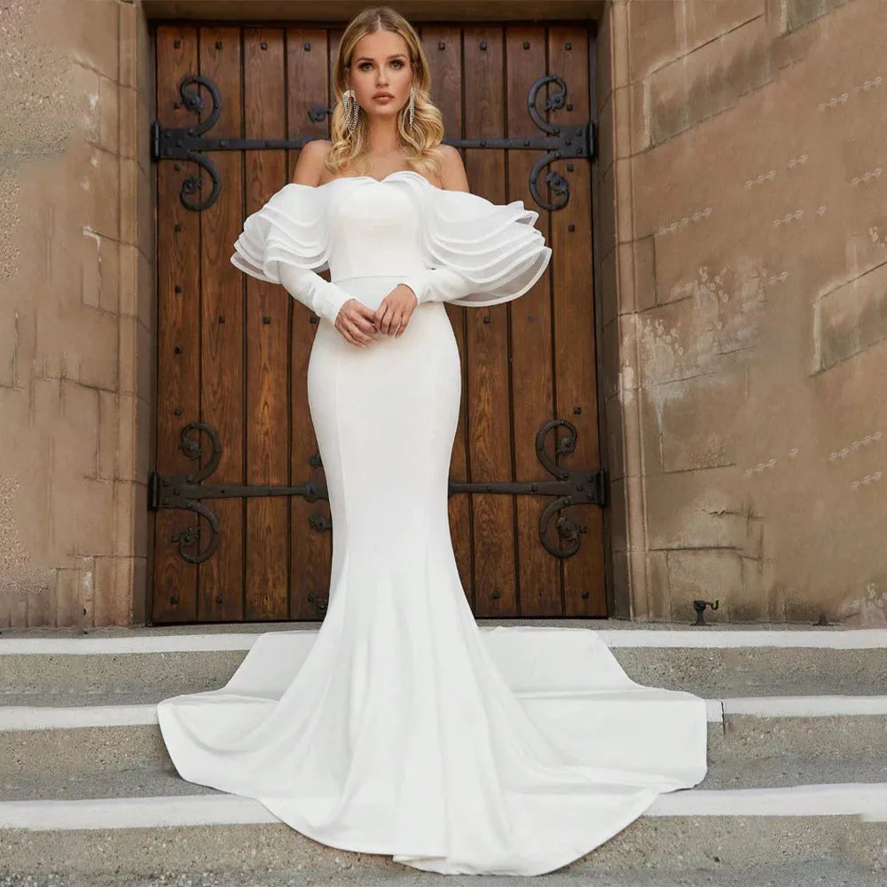 Woman in a fitted off-the-shoulder white gown with dramatic puffed sleeves, standing on stone steps in front of a dark wooden door with ornate hinges.