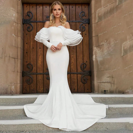 Woman in a fitted off-the-shoulder white gown with dramatic puffed sleeves, standing on stone steps in front of a dark wooden door with ornate hinges.