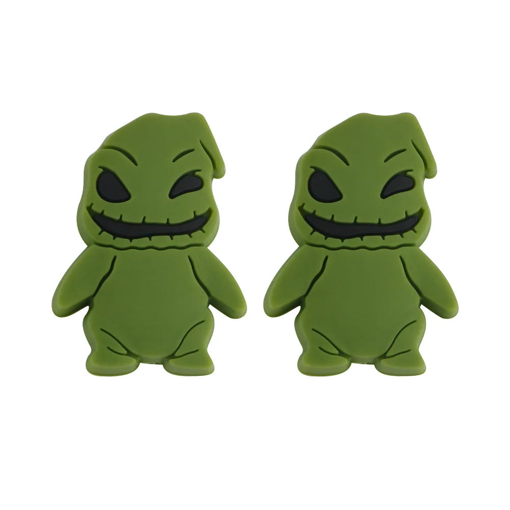 Two identical green cartoon figure earrings with grinning faces and black eyes. They have a textured, rounded shape resembling a playful, spooky character.