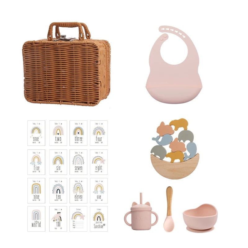 A wicker suitcase, pink bib, stacking toy, milestone cards, and feeding set including a cup, spoon, and bowl. The cards display months one to twelve with rainbows.