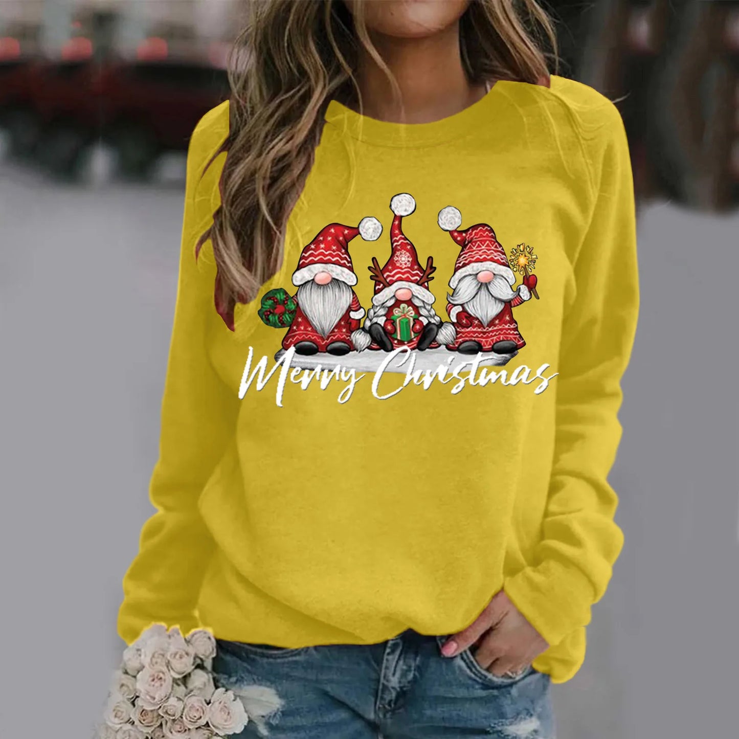 Woman wearing a yellow sweater featuring three festive gnomes in red hats and "Merry Christmas" text. She holds white roses and wears distressed jeans.
