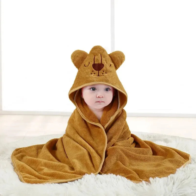 A baby wrapped in a plush, brown hooded towel with bear ears and face embroidery, sitting on a soft white rug in a bright room with a white background.