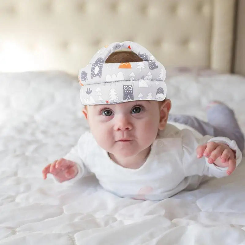 Baby lying on a white bed wearing a padded head protector with cute owl and nature prints, gazing upward. The background features a soft, tufted headboard.