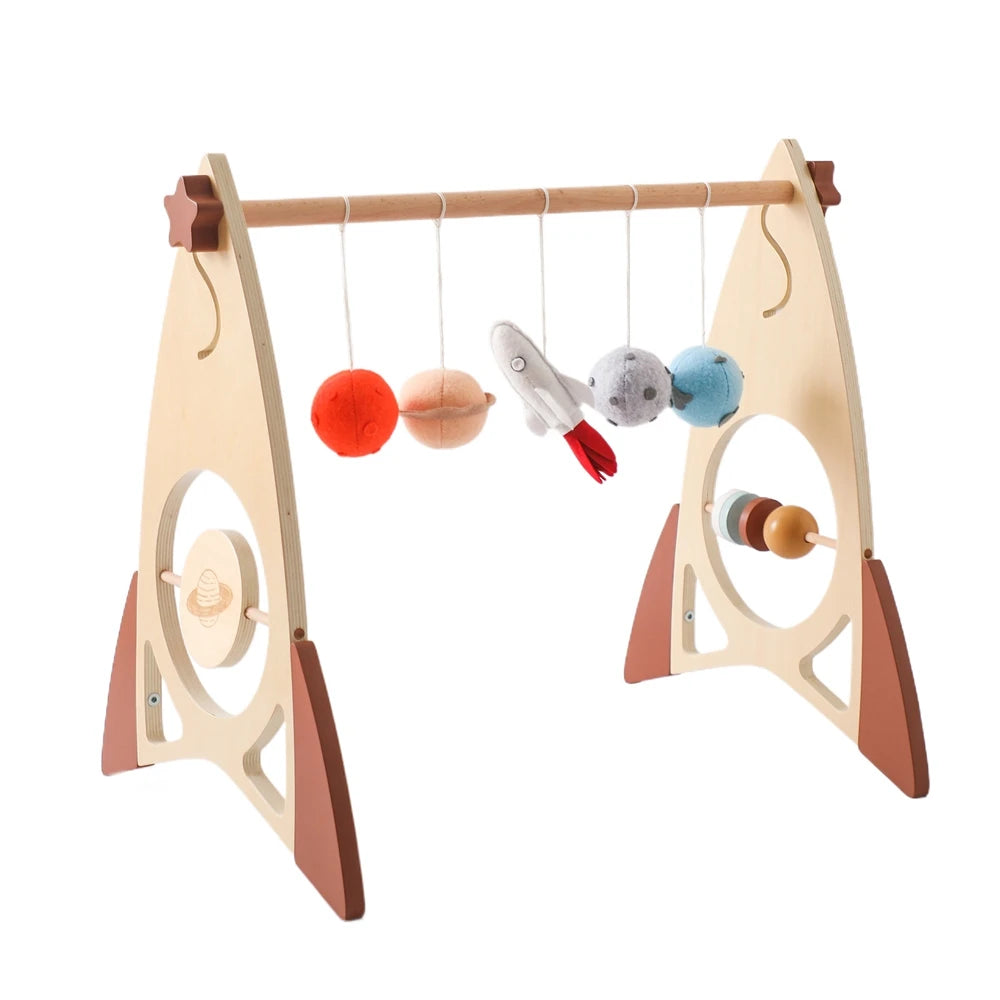 Wooden baby play gym with hanging colorful felt balls, a plush rocket, and geometric shapes. It features a sturdy A-frame design with circular cutouts and rotating elements.