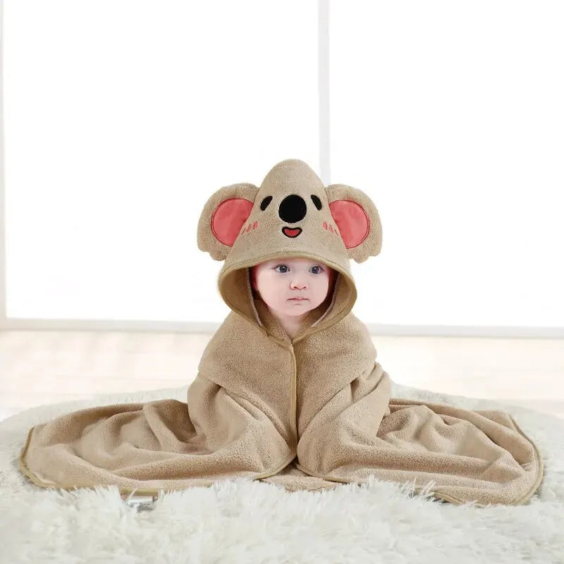 Baby wrapped in a beige koala-themed hooded blanket, sitting on a white fluffy rug against a bright windowed background. The hood features cute koala ears and face.