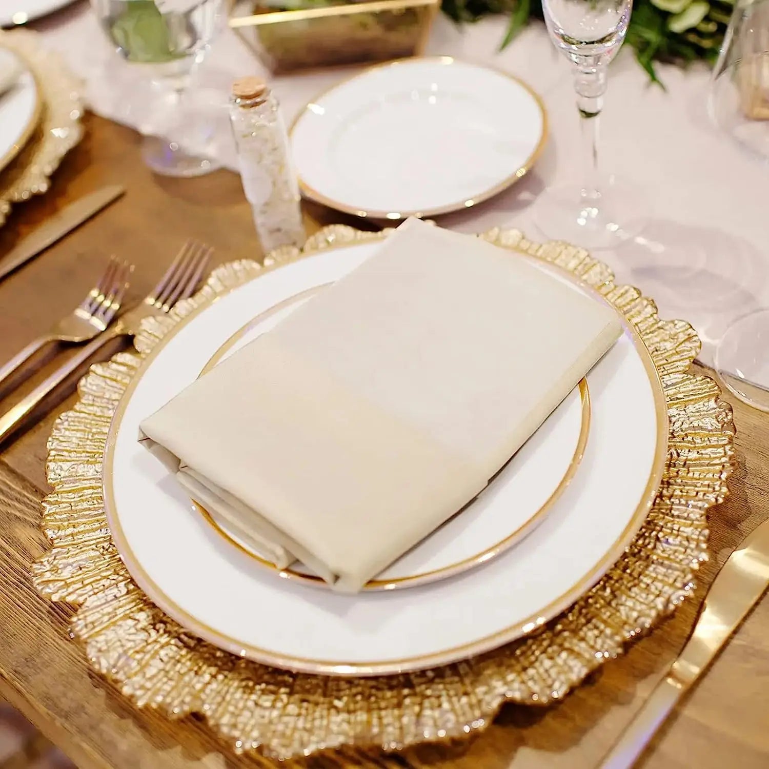 Elegant table setting with gold-rimmed plates, a folded napkin, gold charger, cutlery, wine glass, and decorative greenery on a wooden table.
