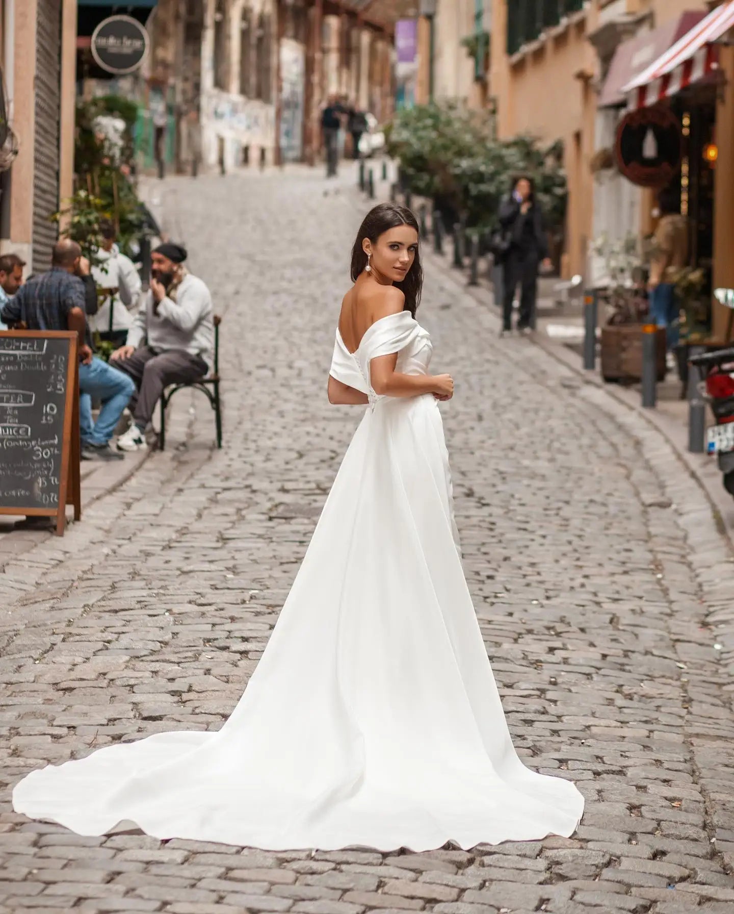 Woman in an elegant off-shoulder white gown stands on a cobblestone street, surrounded by rustic buildings. She looks over her shoulder, with onlookers sitting nearby.