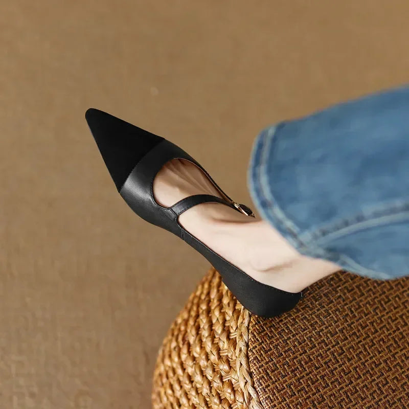 A foot wearing a black pointed-toe, single-strap flat shoe rests on a woven round stool. The person is wearing blue denim jeans, visible against a plain beige background.