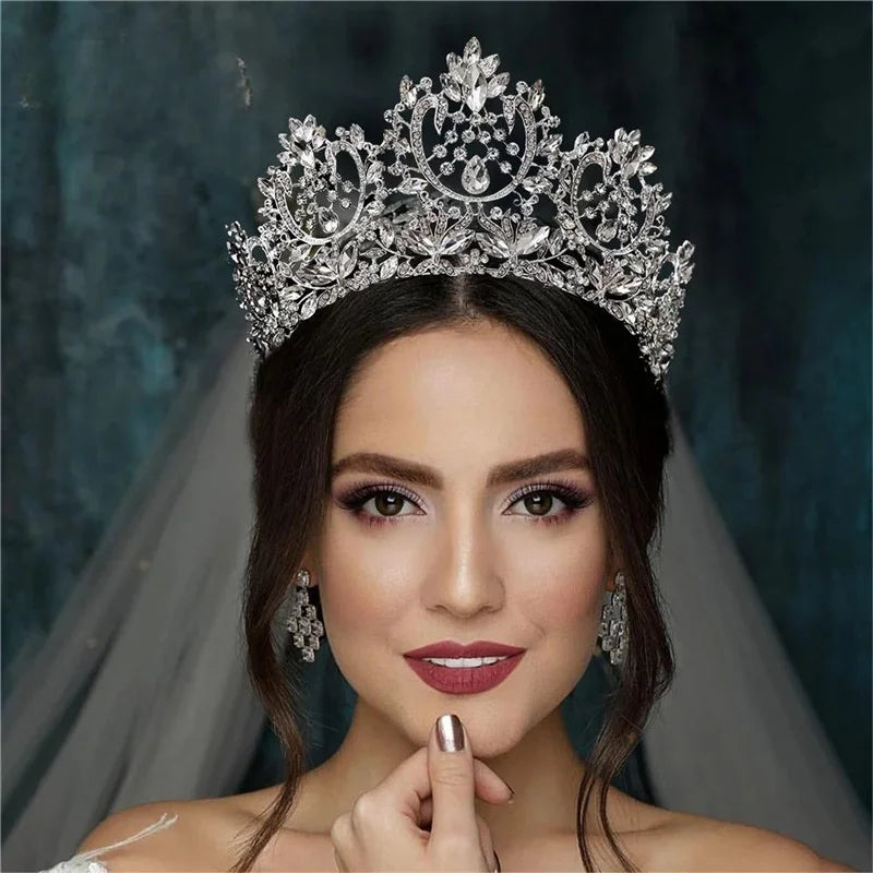 A woman wearing a detailed, silver tiara with intricate designs and matching earrings. She has dark hair, elegant makeup, and a contemplative expression against a dark background.