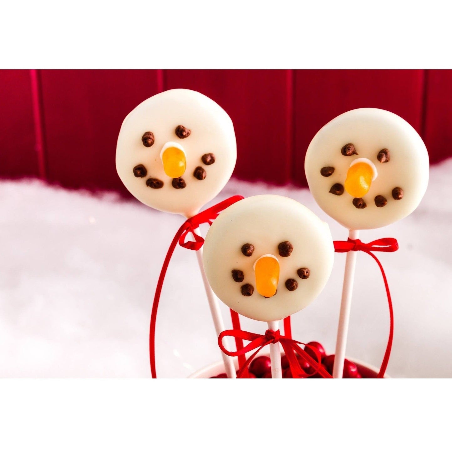 Three snowman-themed cake pops with white icing, chocolate chip eyes, orange candy noses, and red ribbon bows on sticks, set against a snowy backdrop.