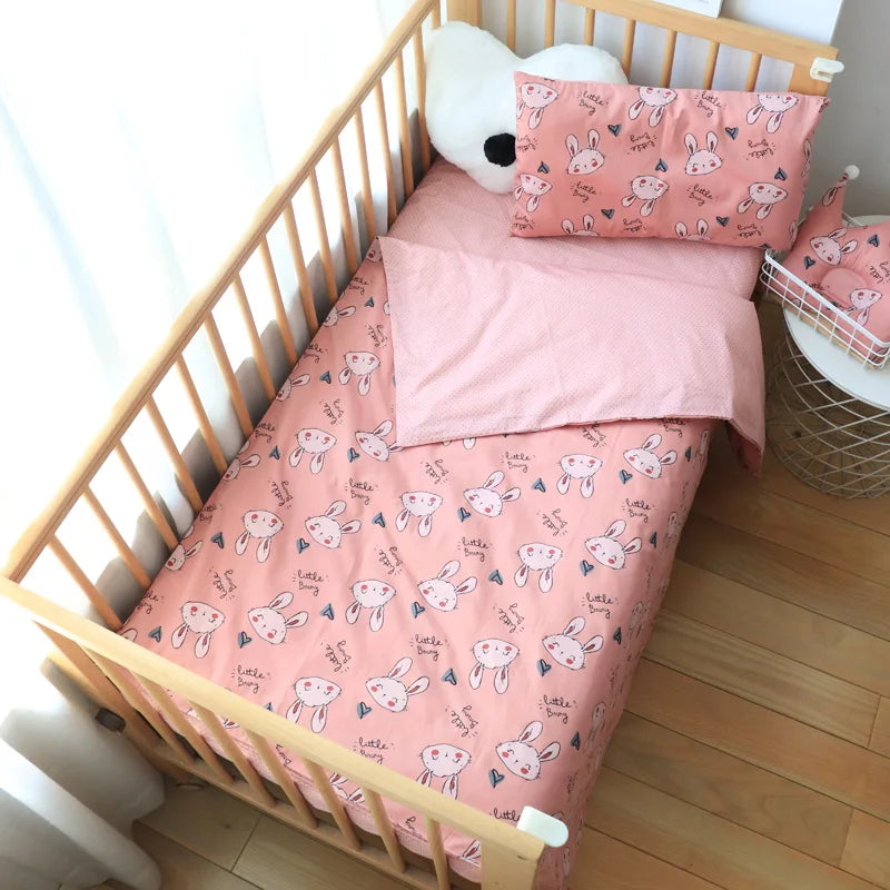 Wooden crib with pink bunny-themed bedding, including a sheet and pillow. A white, bear-shaped cushion rests at the head, with a small round table nearby.