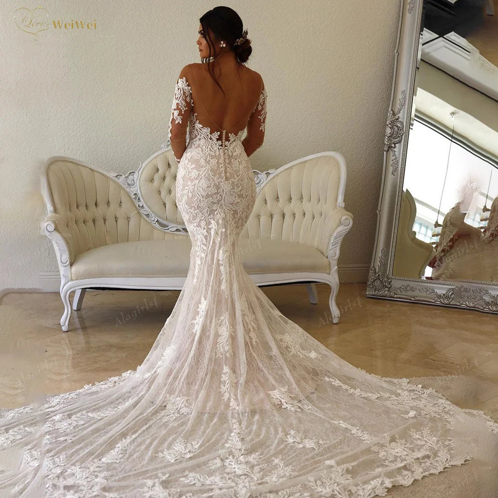 A woman in a lace wedding gown with a long train stands in front of an ornate mirror and vintage sofa, her back facing the camera, in an elegant room.