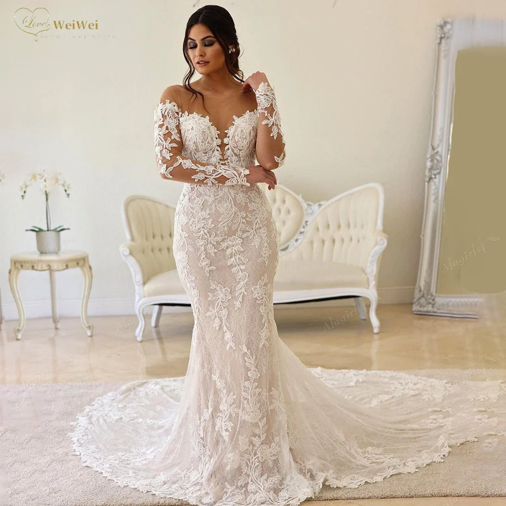 Bride in elegant lace wedding gown with long train stands indoors by vintage sofa; soft lighting enhances floral patterns and off-shoulder design.