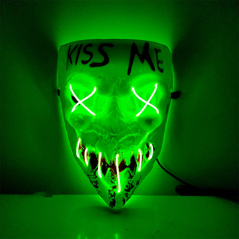 A neon green glowing mask with "KISS ME" written on the forehead, featuring X-shaped eyes and pointed teeth, hanging on a wall.