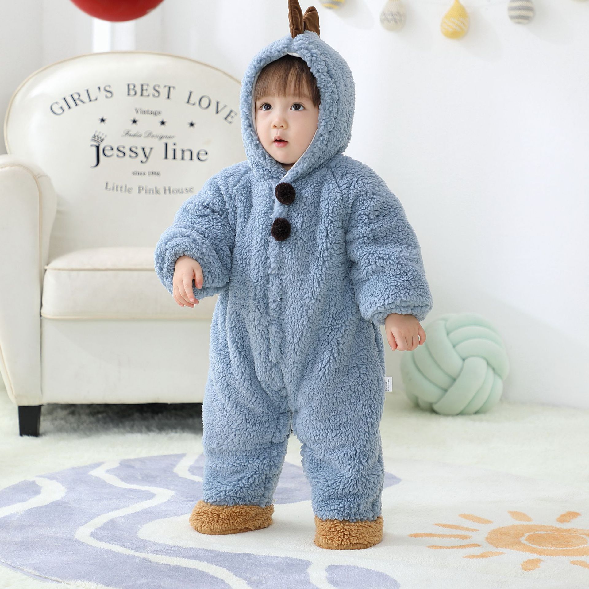 A toddler in a blue fuzzy onesie stands on a patterned rug in a cozy room, next to a white armchair. The outfit features bear-like ears and brown paw-shaped feet.