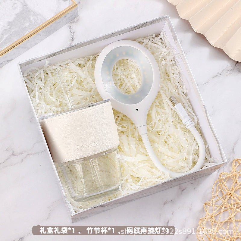 A gift box contains a stylish glass cup with a beige sleeve and a white circular USB LED lamp, both nestled in shredded paper on a marble surface.