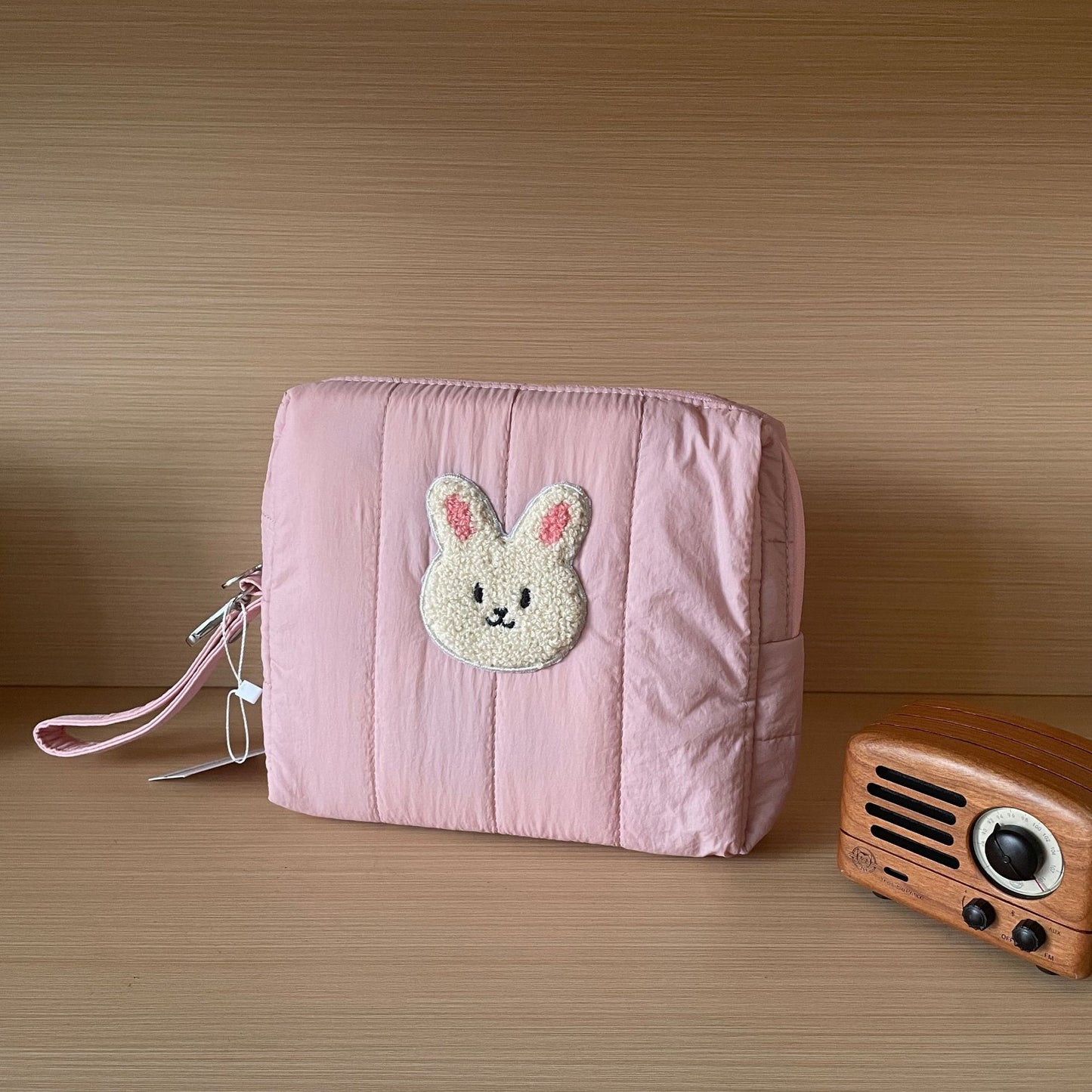 A pink quilted pouch with a cute bunny applique on the front sits on a wooden surface. A vintage-style wooden radio is placed beside it, against a light beige background.