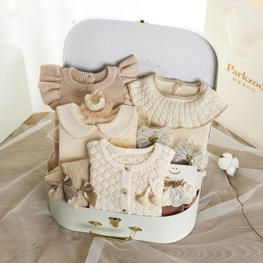 A collection of baby clothes in soft pastel shades, neatly arranged in a white suitcase. The set includes knitted sweaters, delicate bows, and lace details, placed on a wooden surface.