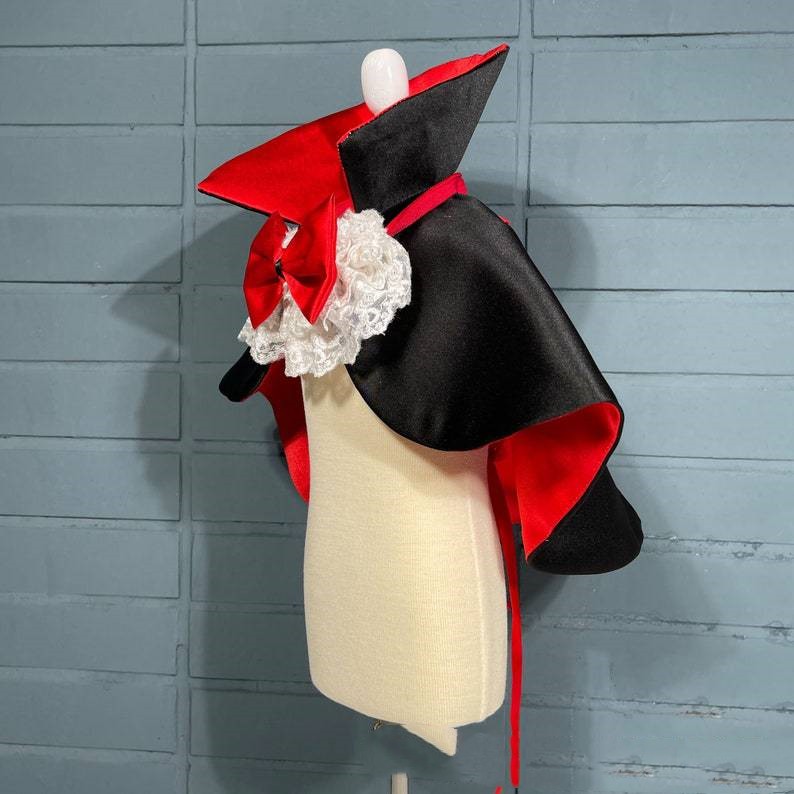 A mannequin displays a black and red vampire cape with a dramatic collar and white lace detailing against a light blue background.