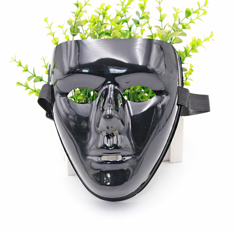A glossy black face mask with cutout eyes and mouth is displayed with green foliage in the background. The mask has a black strap on the sides for wearing.