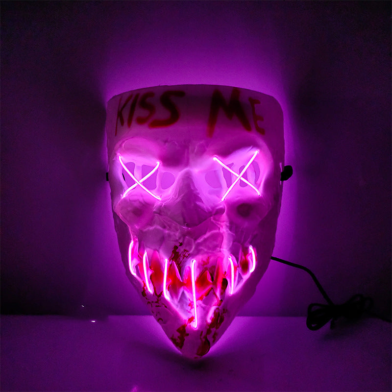 A white mask with pink neon lights forming an "X" over the eyes and sharp teeth. The phrase "Kiss Me" is written on the forehead in red. Black cord visible on the side.