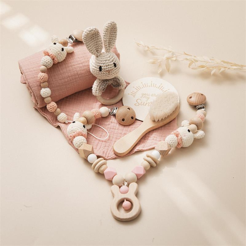 Crocheted bunny toy, baby brush, wooden rattle, and pacifier clip with beads on a pink fabric. Nearby, a decorative plaque reads "you are my sunshine" with dried leaves.