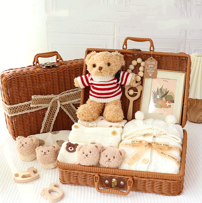Wicker baby gift set with a teddy bear in a striped sweater, framed picture, knit hat, teddy face slippers, plush toys, and a teether. The case has lace accents.
