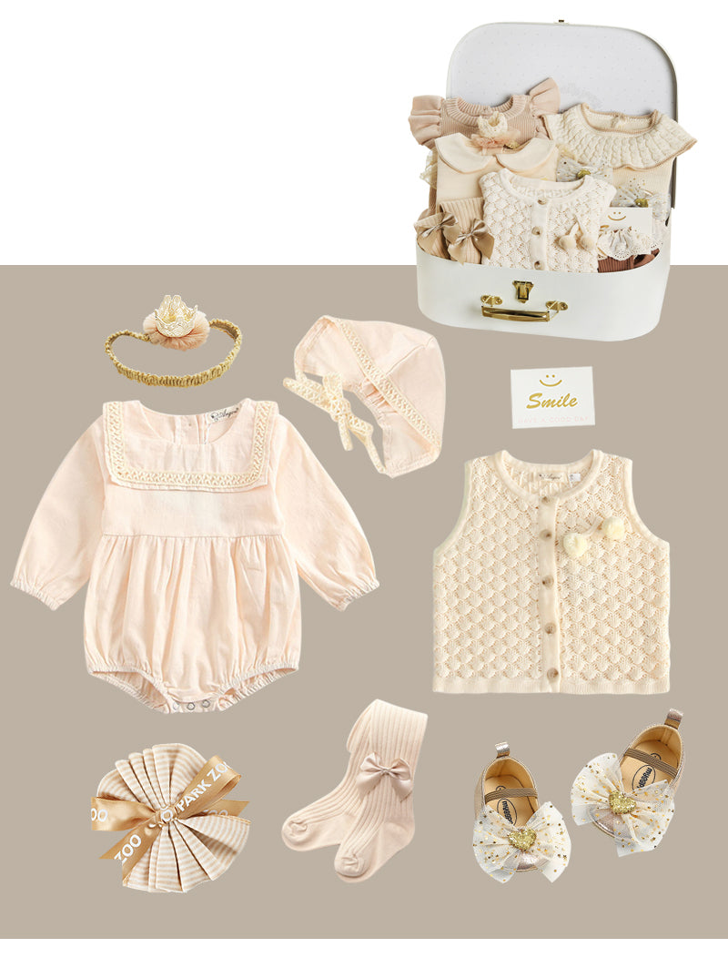 Baby clothing set in light pink and cream, featuring a long-sleeved romper, knit vest, beanie, headband, socks, and shoes with bows, displayed with a small suitcase and card.