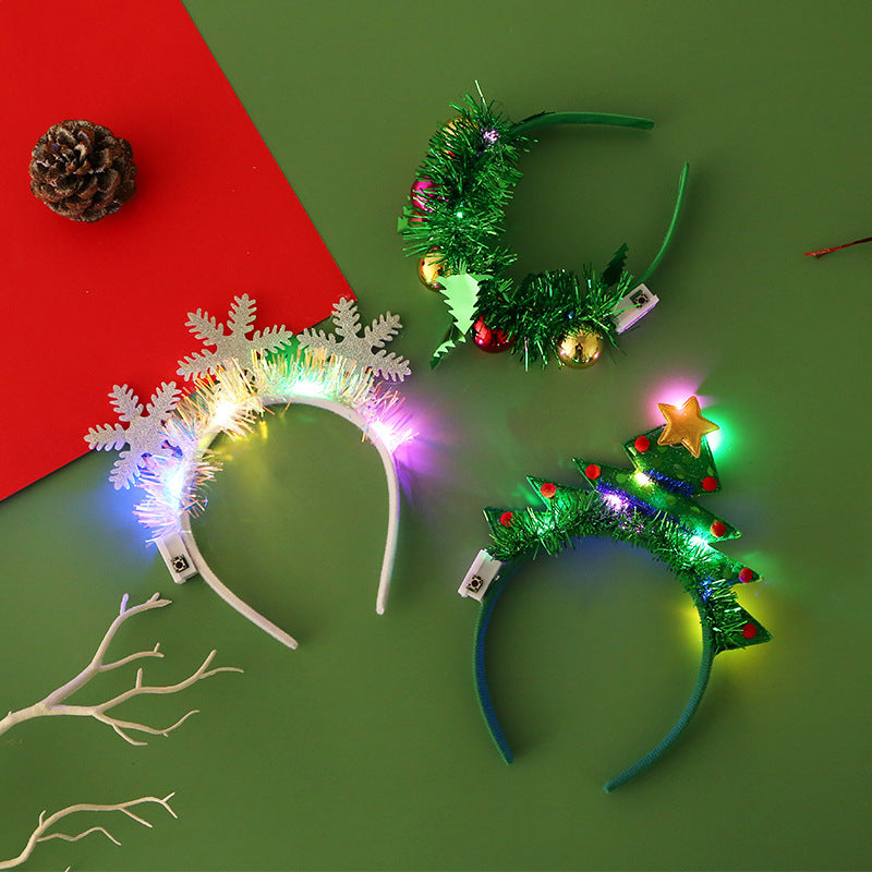 Three festive headbands on a green background: a snowflake with lights, a tinsel wreath with baubles, and a Christmas tree with a star. Pinecone and red paper nearby.