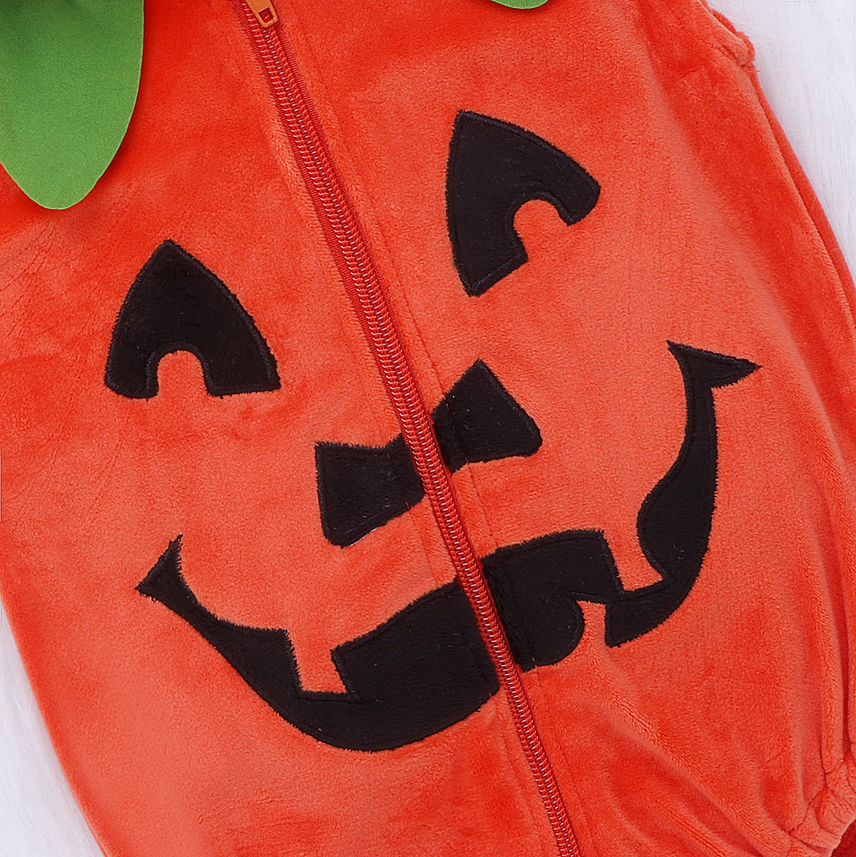 Close-up of a fuzzy orange baby outfit with a zipper, featuring a black cartoon jack-o'-lantern face and green felt leaves on the hood.