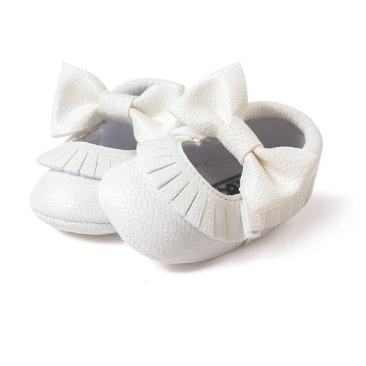 White baby shoes made of textured material, featuring large bows and fringe detailing. The design is elegant and suitable for special occasions or casual wear.