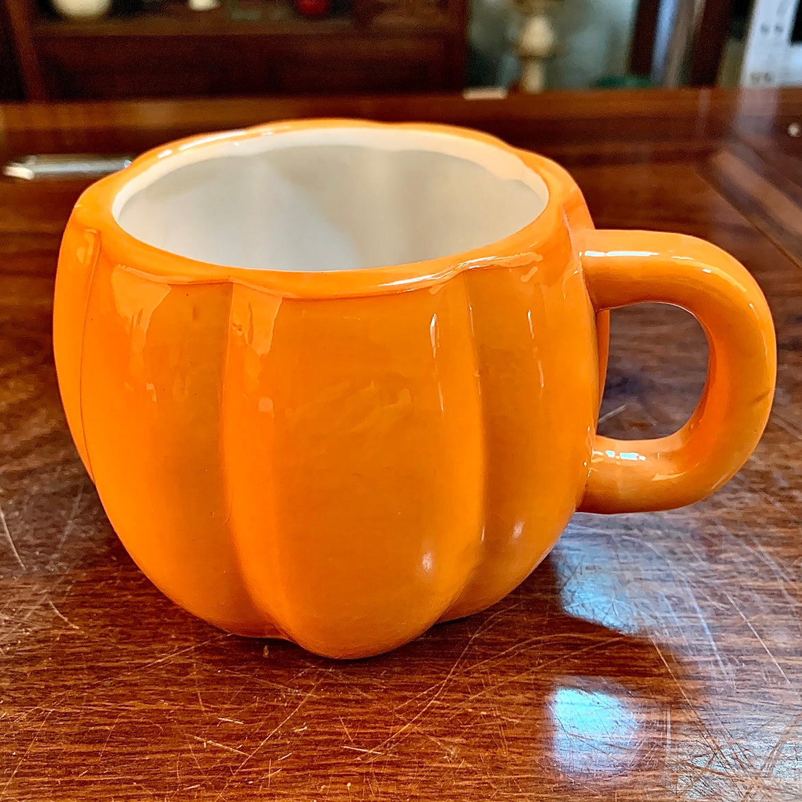 Orange mug shaped like a pumpkin with a glossy finish sits on a wooden table. The mug has a curved handle and a smooth, light-colored interior.