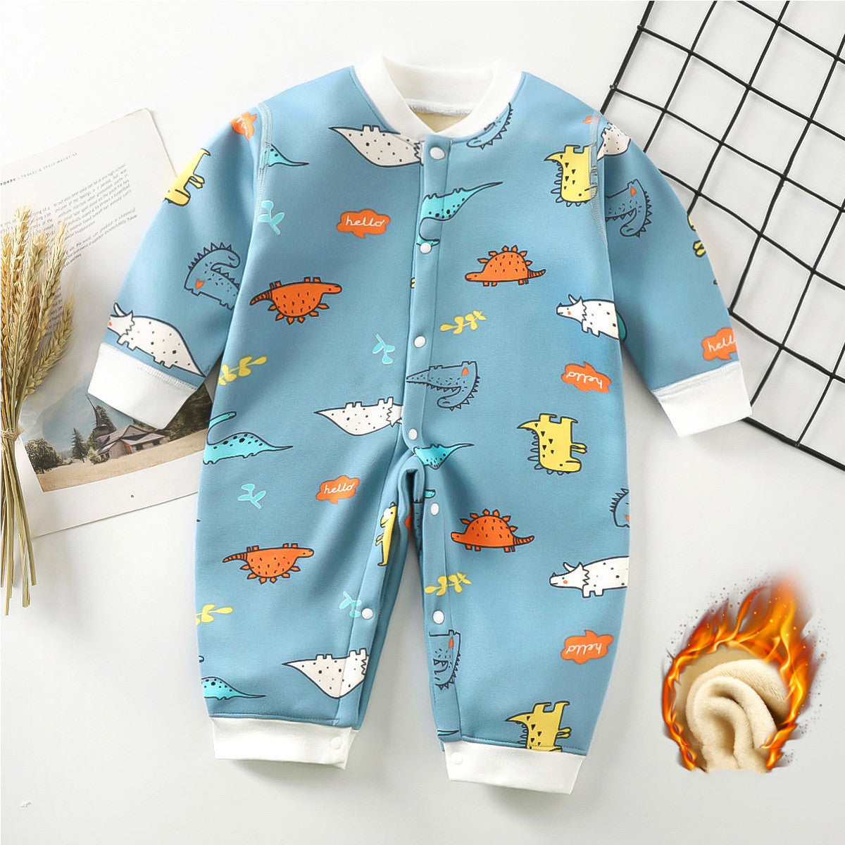 Blue baby jumpsuit with colorful dinosaur patterns and "hello" text, long sleeves, and snap buttons. White cuffs and collar. Dried plants and a grid pattern decorate the background.