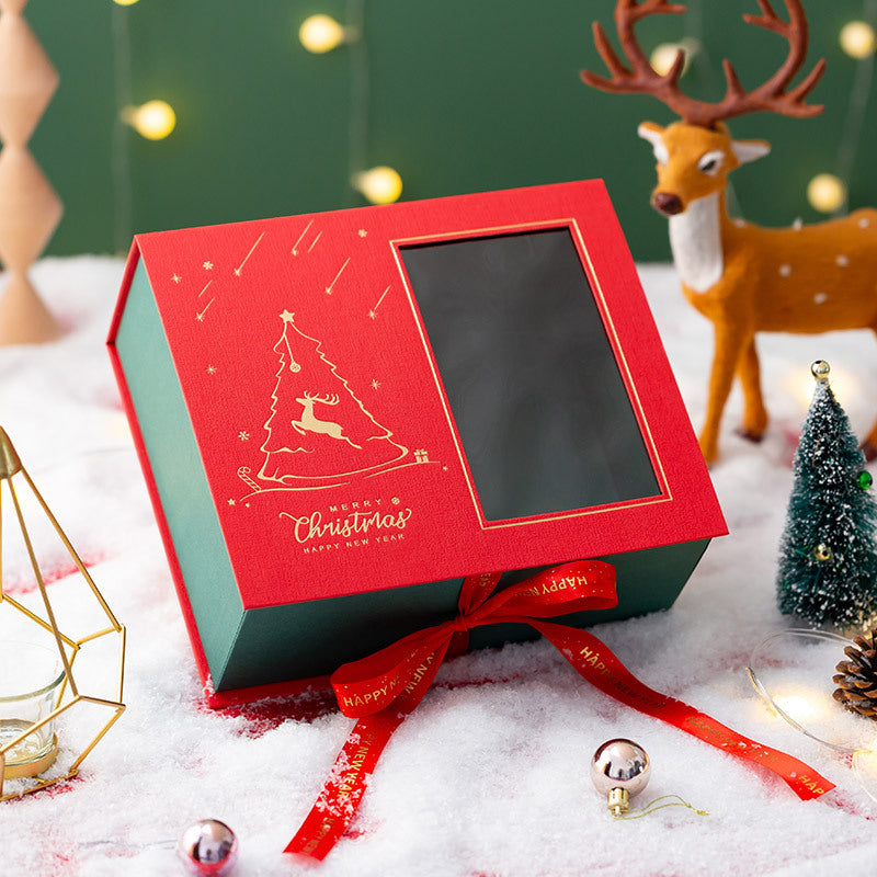 Red and green Christmas gift box with a reindeer design, tied with a red ribbon, surrounded by snow, fairy lights, a deer figurine, and small Christmas trees.