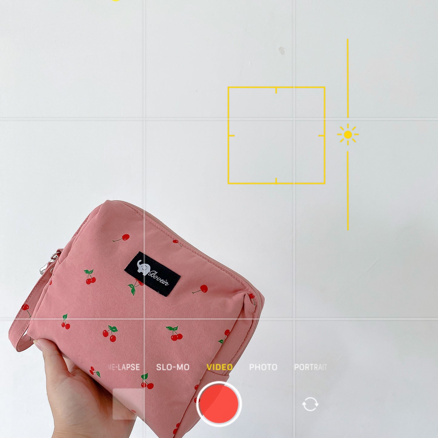 A hand holds a pink pouch with cherry prints against a white background. The phone camera interface is visible, set to video mode, with on-screen controls and yellow focus grid.