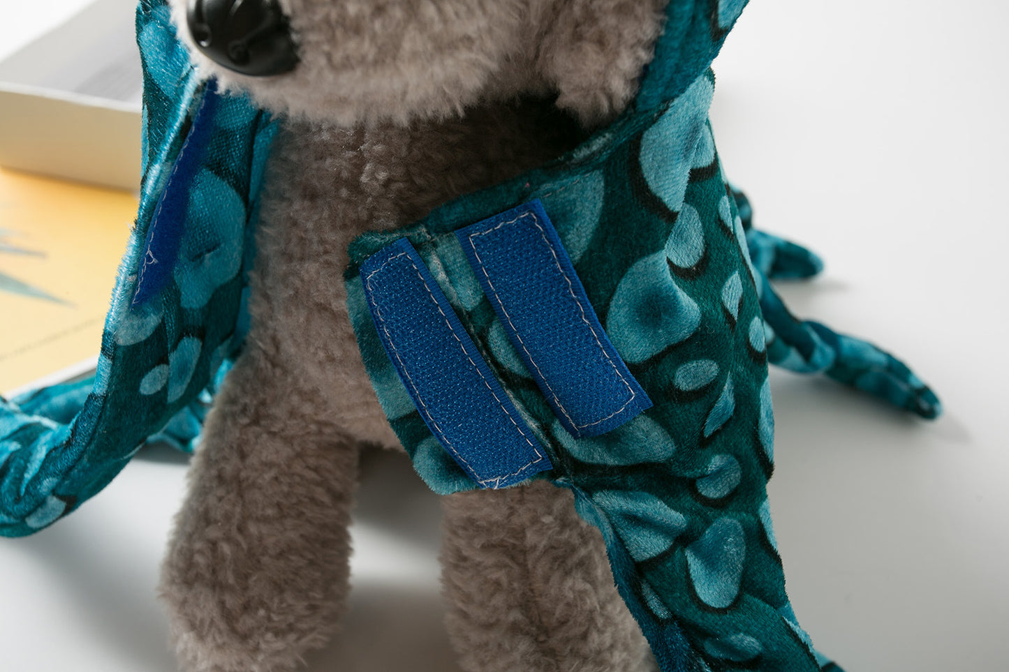 A close-up of a gray plush toy wearing a teal floral-patterned cape with blue Velcro fasteners. The toy is sitting on a white surface beside an open book.