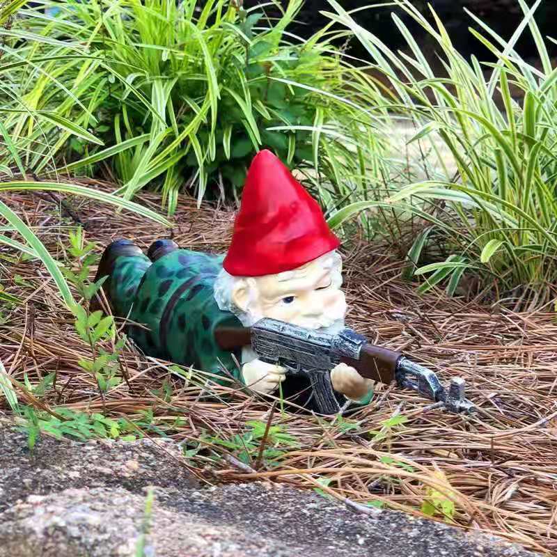 Garden gnome in a green outfit and red hat, lying on pine needles, playfully aiming a toy rifle, surrounded by lush greenery.