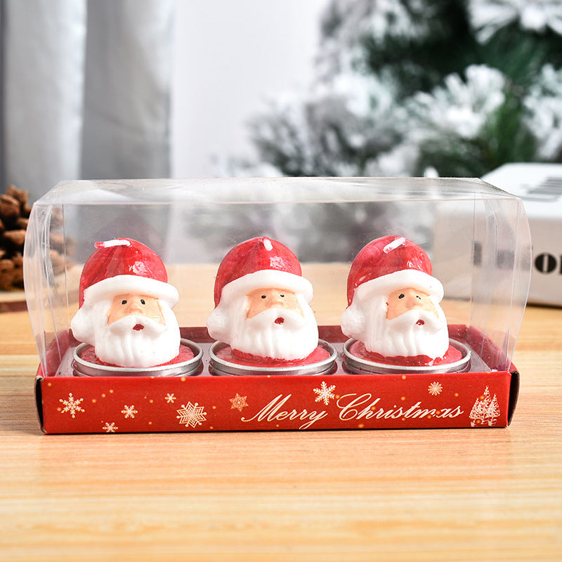 Three Santa Claus-shaped candles in a red "Merry Christmas" box, each with a white beard and red hat. They are encased in a clear plastic cover on a wooden surface.
