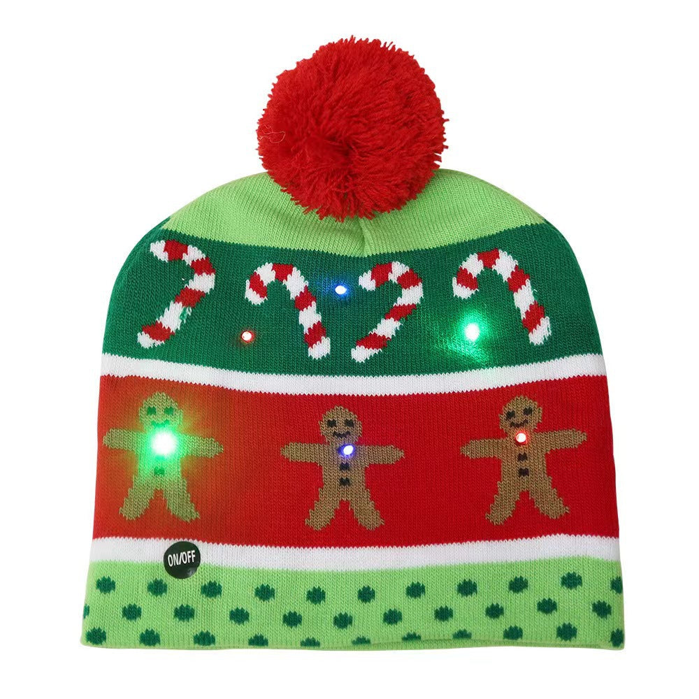 Green and red beanie with a pompom, featuring candy cane and gingerbread men patterns. The hat has colorful LED lights and an on/off button, suitable for festive occasions.