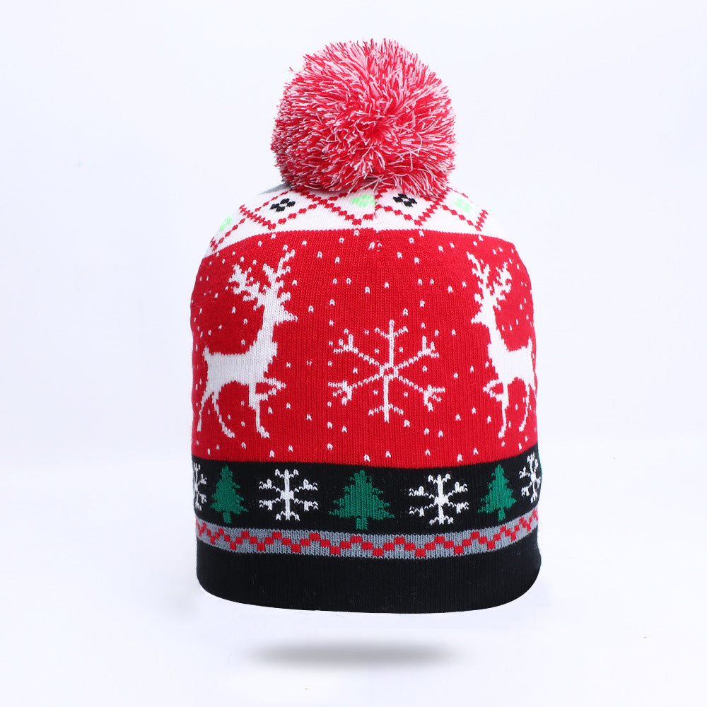 Red winter beanie with white reindeer, snowflakes, and tree designs, topped with a red and white pom-pom. The lower band features a black background with festive patterns.