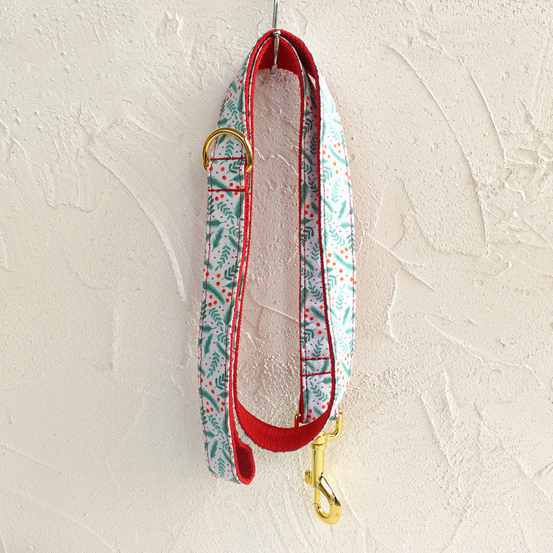 A colorful dog leash with a festive pattern of green leaves and red dots hangs against a textured white wall. The leash features a gold clasp and red edging.