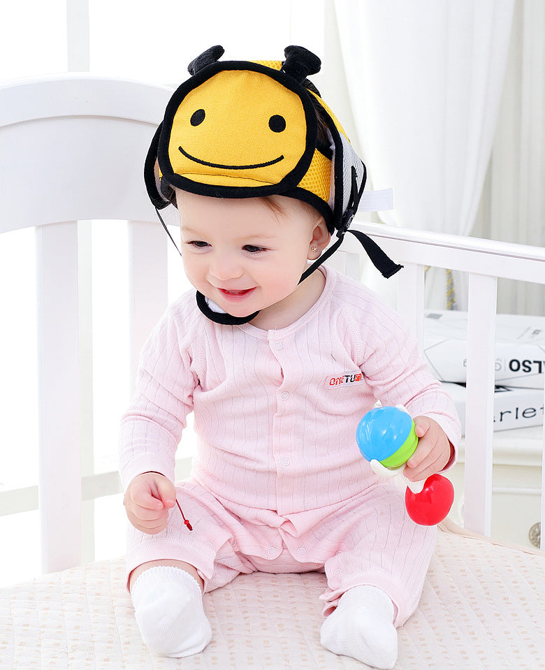 A baby in a pink onesie sits in a white crib, smiling while wearing a cute bee-themed helmet. The baby holds a colorful toy rattle and looks content.