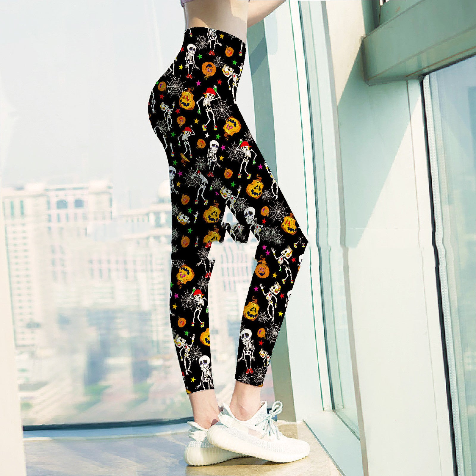 A person stands indoors wearing black leggings decorated with skeletons, pumpkins, and stars. They are wearing white sneakers and are beside large windows with a city view.