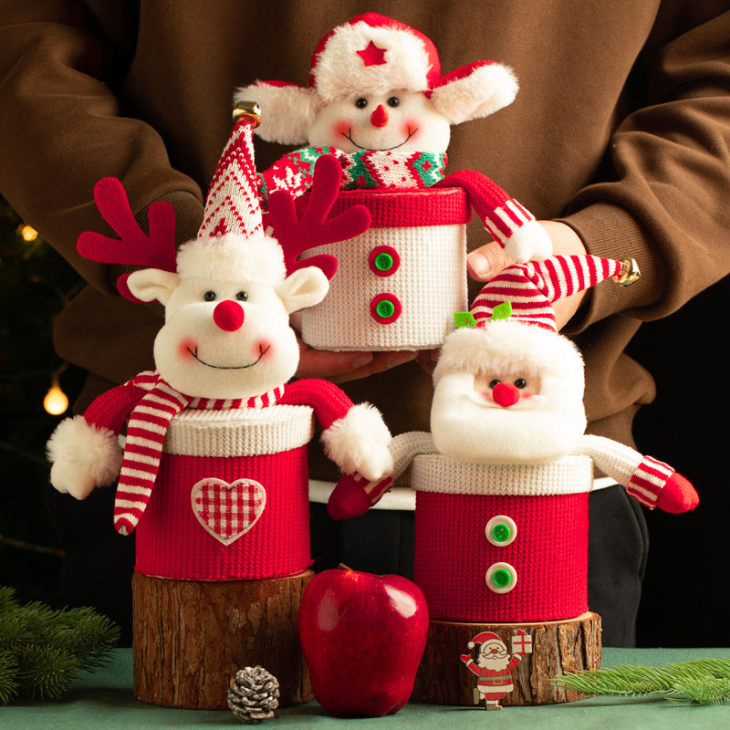 Festive plush toys, including a reindeer, Santa, and a snowman, in red and white knit outfits with buttons. They rest on wood slices. A red apple and pine cone sit in front.
