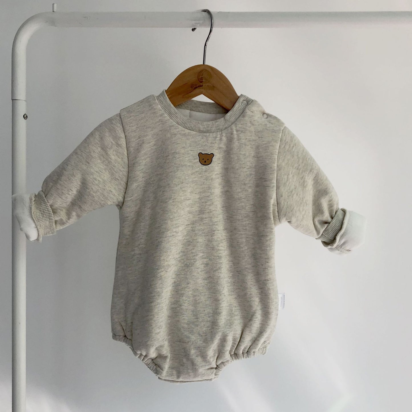 A light gray baby onesie with rolled-up sleeves hangs on a wooden hanger. It features a small brown bear patch on the chest, displayed against a plain white background.