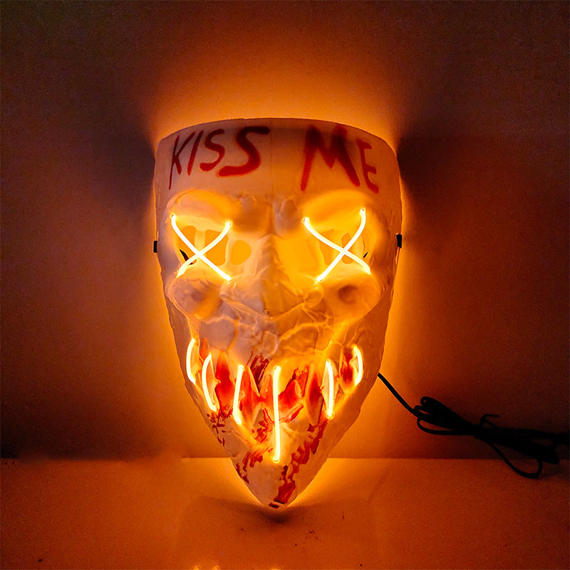 Glowing Halloween mask with neon details, featuring "Kiss Me" in red, crossed-out eyes, and jagged teeth. The eerie lighting creates a sinister atmosphere.